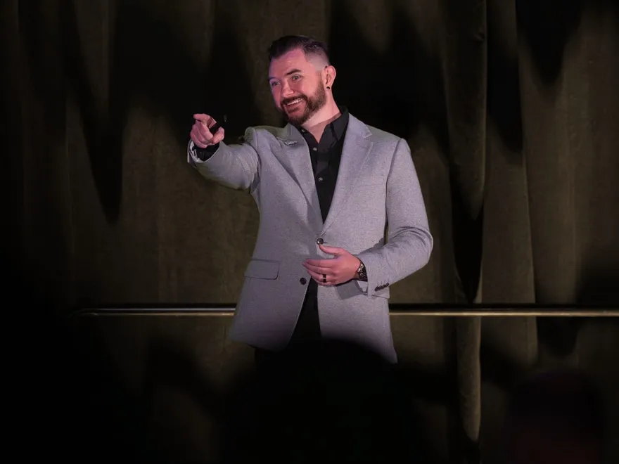 Kyle Christensen on stage in a gray blazer, smiling and pointing with his right arm, in front of a dark curtain